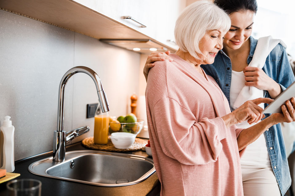 A grandmother and her daughter looking at a tablet while standing next to the sink A grandmother and her daughter looking at a tablet while standing next to the sink